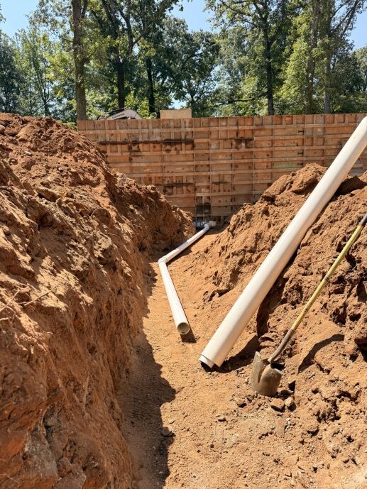 A narrow dirt trench containing white PVC pipes for a waterproofing service, with a shovel leaning against the side and a tall concrete formwork wall in the background.